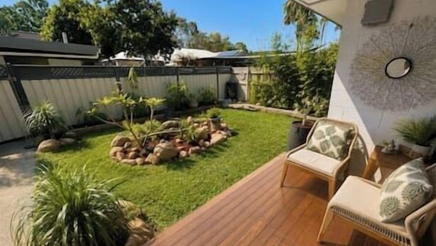 Um quintal com um deck de madeira, área de estar com almofadas, pequeno jardim com pedras e plantas, e um objeto decorativo fixado na parede.