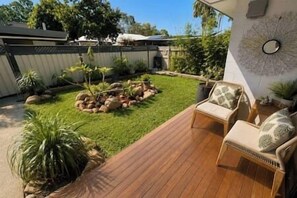 A backyard with a wooden deck, a seating area with cushions, a small garden with rocks and plants, and a wall-mounted decorative piece. - Coastal Hideaway at Kissing Point | Tropical 2BR Unit (North Ward)
