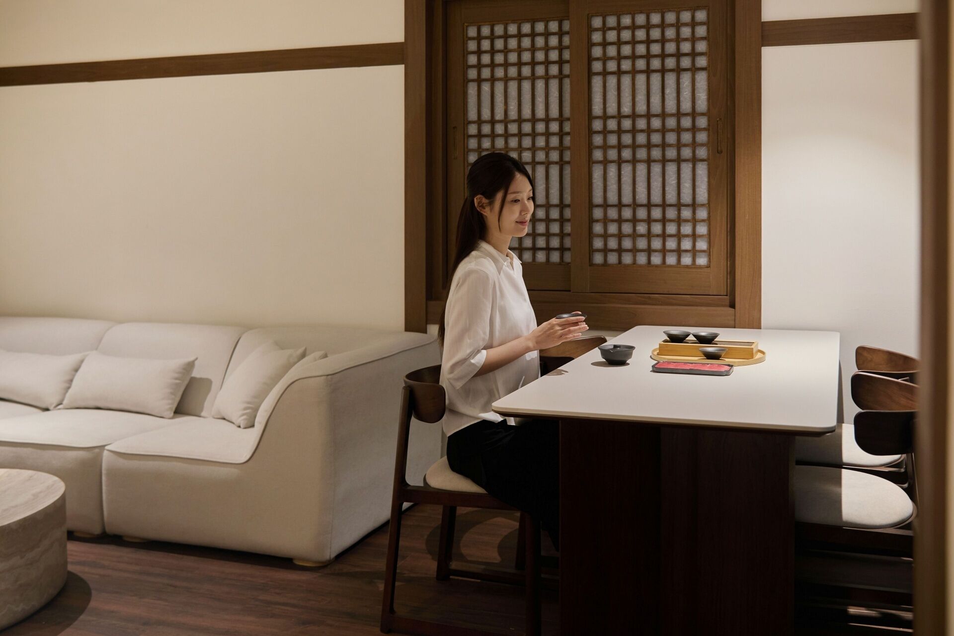 A person seated at a table with a traditional tea set, a sofa, and a wooden partition in the background.