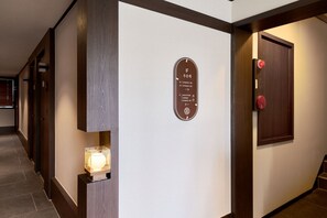 Un pasillo con una recepción, un cartel en la pared con los números de las habitaciones y una puerta con pomo rojo.
