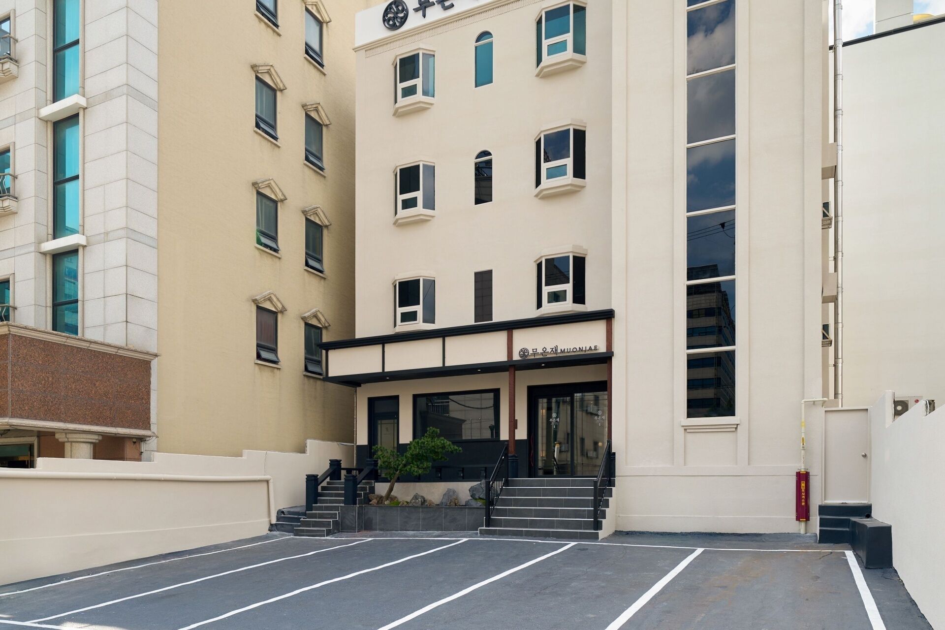 Un edificio de varios pisos con entrada a nivel de calle, una escalera que conduce a la entrada y un área de estacionamiento al frente.