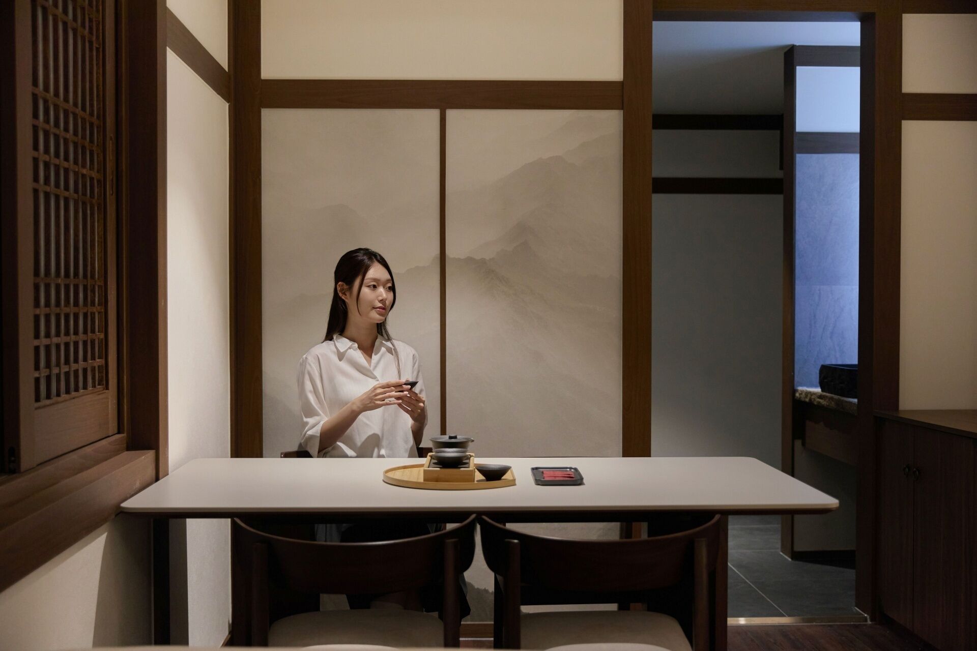 A person seated at a table with a traditional tea set, in a room with sliding doors and a view of a mountain.