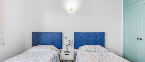 Een slaapkamer met twee enkele bedden, een nachtkastje met een lamp, een witte muur en een blauwgroene deur.