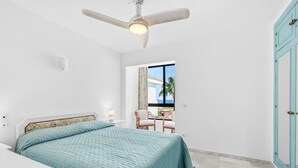 Quarto com cama, ventoinha no teto, mesa de cabeceira, cadeira e janela com vista para palmeiras.