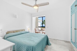 Ein Schlafzimmer mit Bett, Deckenventilator, Nachttisch, Sessel und Fenster mit Blick auf Palmen.