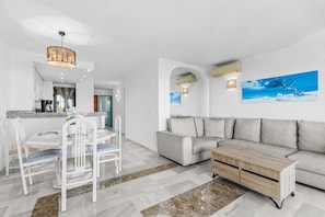 Ein modernes Wohnzimmer mit einer Couch, einem Esstisch mit Stühlen und einer Küchenzeile im Hintergrund.