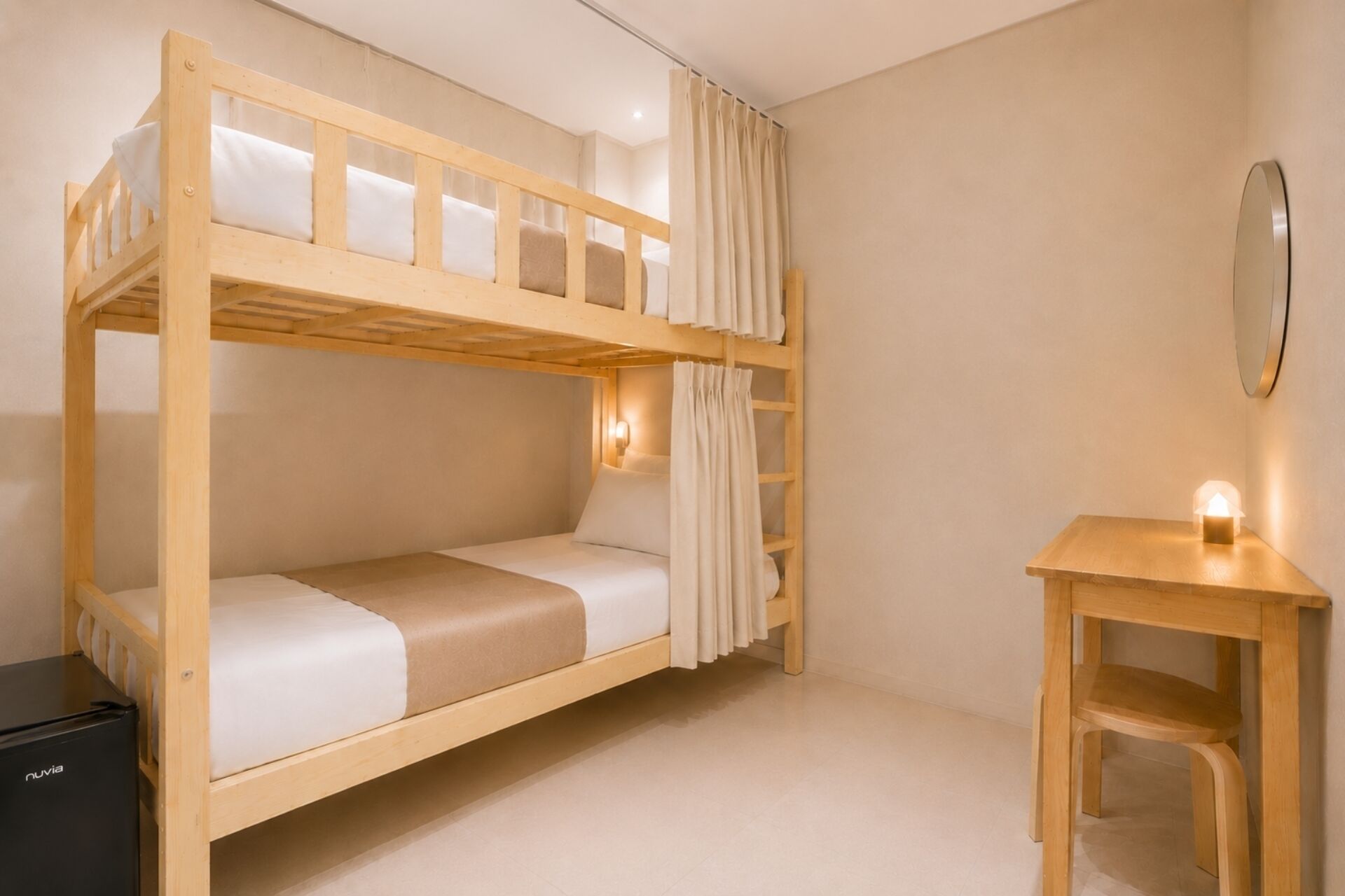 A bunk bed room with a desk and a small table, a wall-mounted mirror, and a black appliance.
