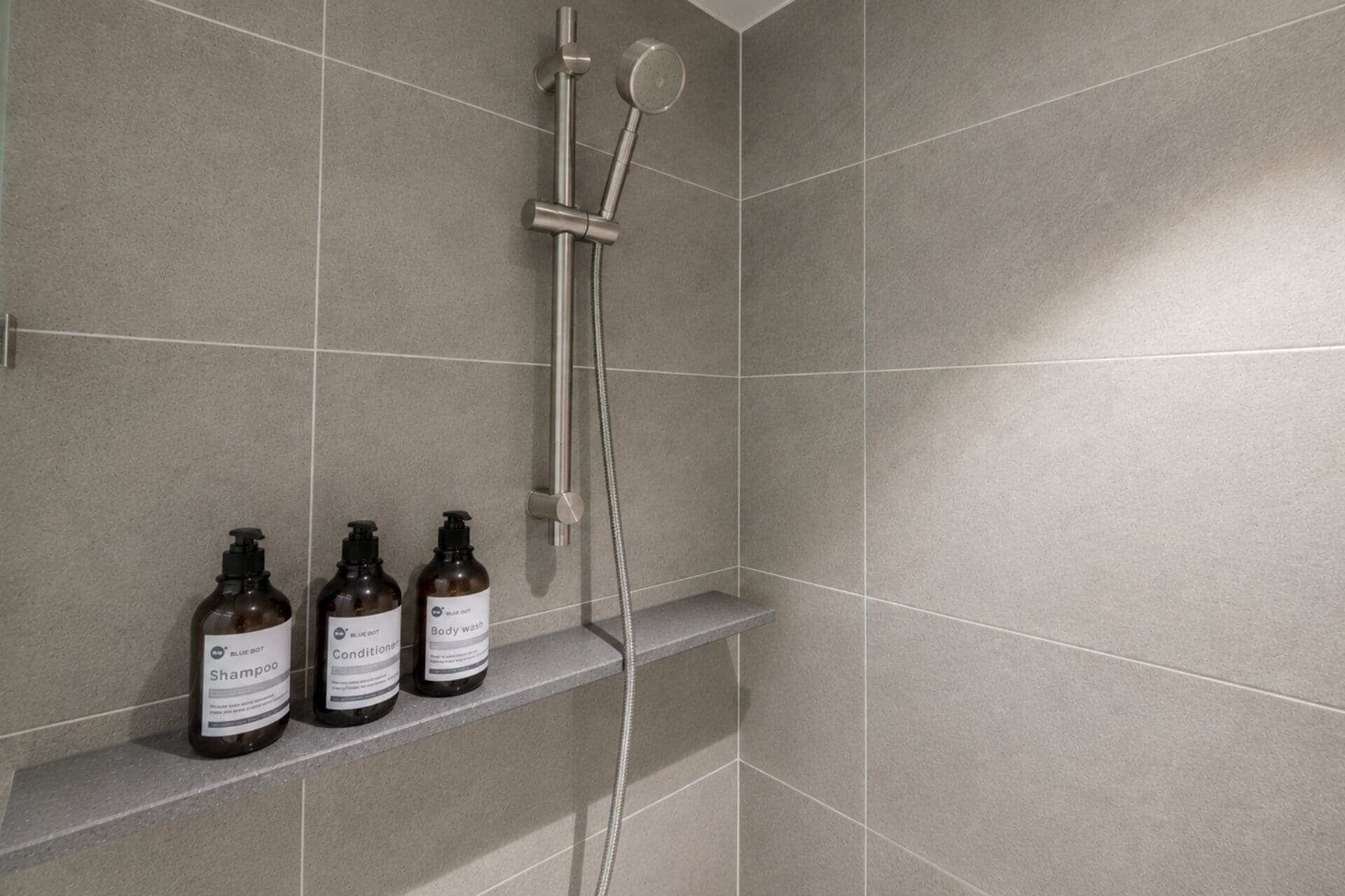 Un espace douche avec des carreaux gris, une douchette et trois bouteilles de produits de la marque Blue Dot, disposées sur une étagère.