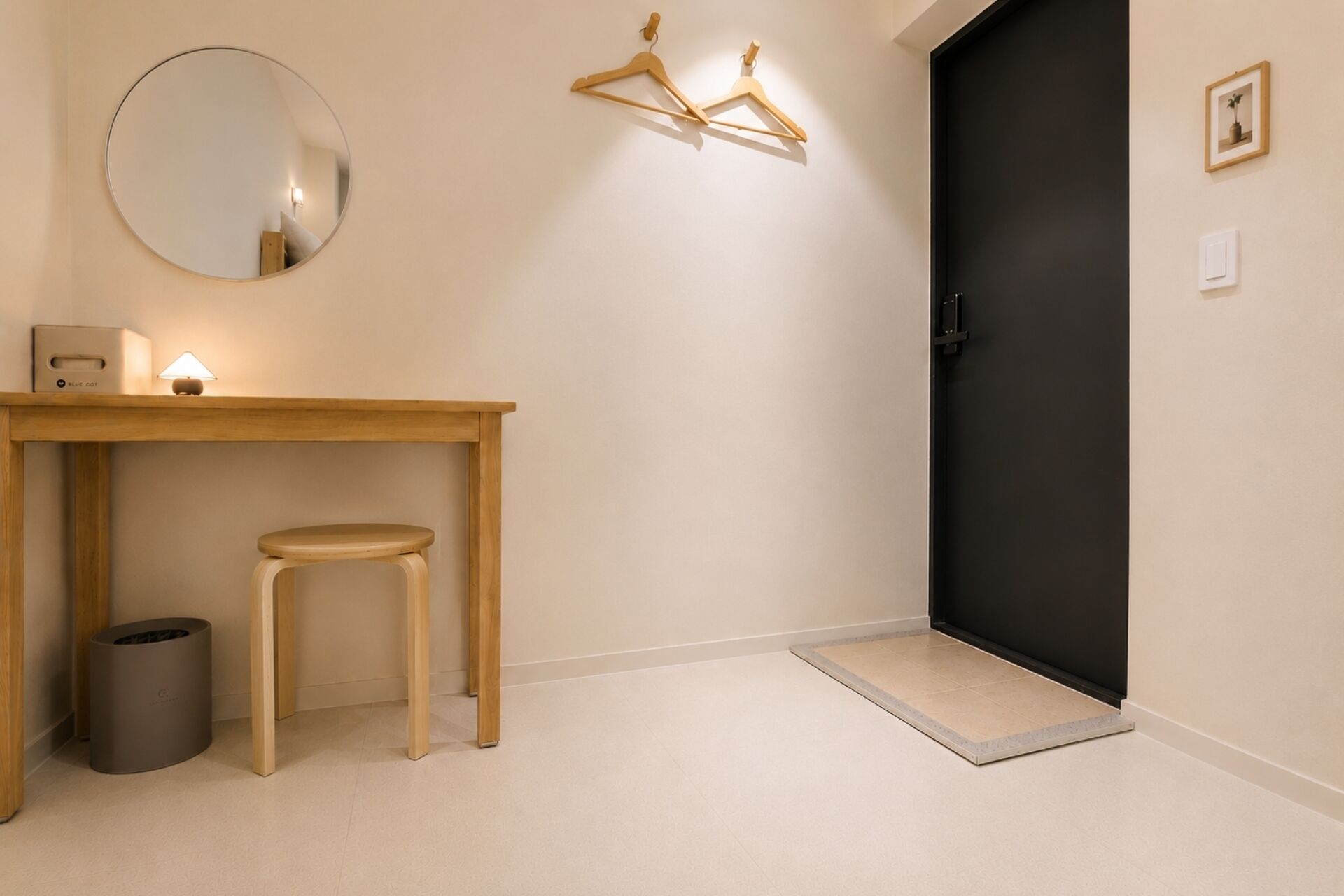 Un pasillo minimalista con una mesa de madera, un taburete, un bote de basura, una puerta negra y un cuadro pequeño en la pared.