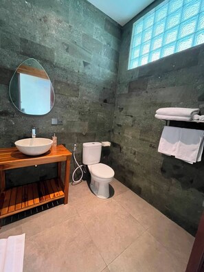 A bathroom with a stone wall, a wooden vanity with a bowl sink, a toilet, and a towel rack with towels.