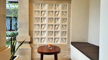 Una pequeña mesa redonda de madera con cojín, ubicada en un rincón junto a un panel decorativo y una planta en maceta a la izquierda.