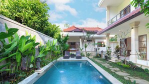 Una piscina con borde de piedra, rodeada de exuberante vegetación y un patio con mobiliario exterior.