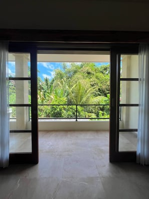 A room with a large sliding glass door leading to a balcony with a view of lush greenery and a clear sky.