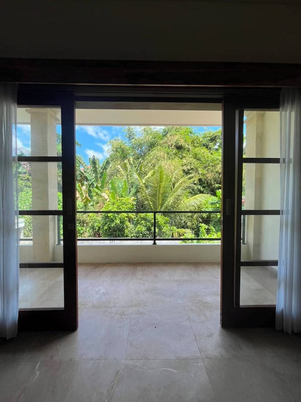 A room with a large sliding glass door leading to a balcony with a view of lush greenery and a clear sky.