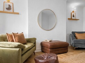 Ein Wohnzimmer mit einem grünen Sofa, einem braunen Hocker, einem grauen Sofa und einem runden Spiegel an der Wand.