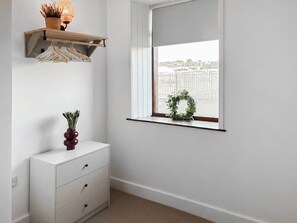 Ein Zimmer mit weißem Schminktisch, einem Holzregal mit Kleidern und einem Fenster mit Blick auf einen Zaun, sowie einem Kranz auf der Fensterbank.