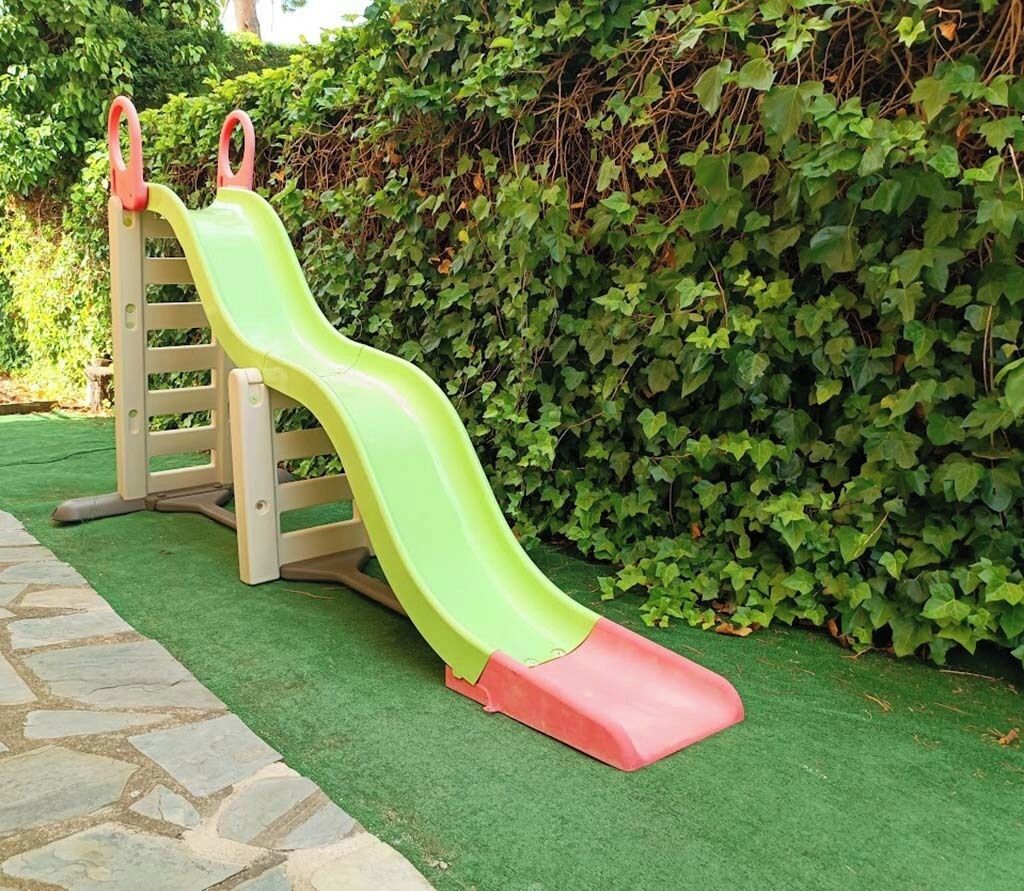 A children's slide with a green and red track, set against a backdrop of green foliage.