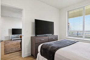 Ein Schlafzimmer mit einem Bett, einem Fernseher auf einer Kommode, einem Fenster mit Jalousien und einer Tür zu einem anderen Raum.