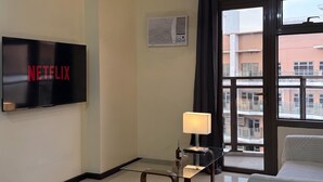 A hotel room with a flat-screen TV displaying the Netflix logo, a standing lamp, a window with a view of buildings, and a bed.