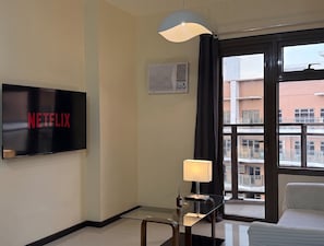 A hotel room with a flat-screen TV displaying the Netflix logo, a standing lamp, a window with a view of buildings, and a bed.