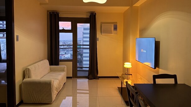 A hotel room with a sofa, a dining table, a TV, and a view of the cityscape through the window.