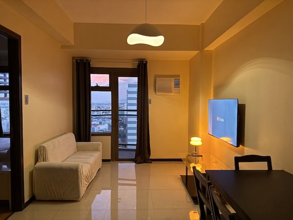 A hotel room with a sofa, a dining table, a TV, and a view of the cityscape through the window.