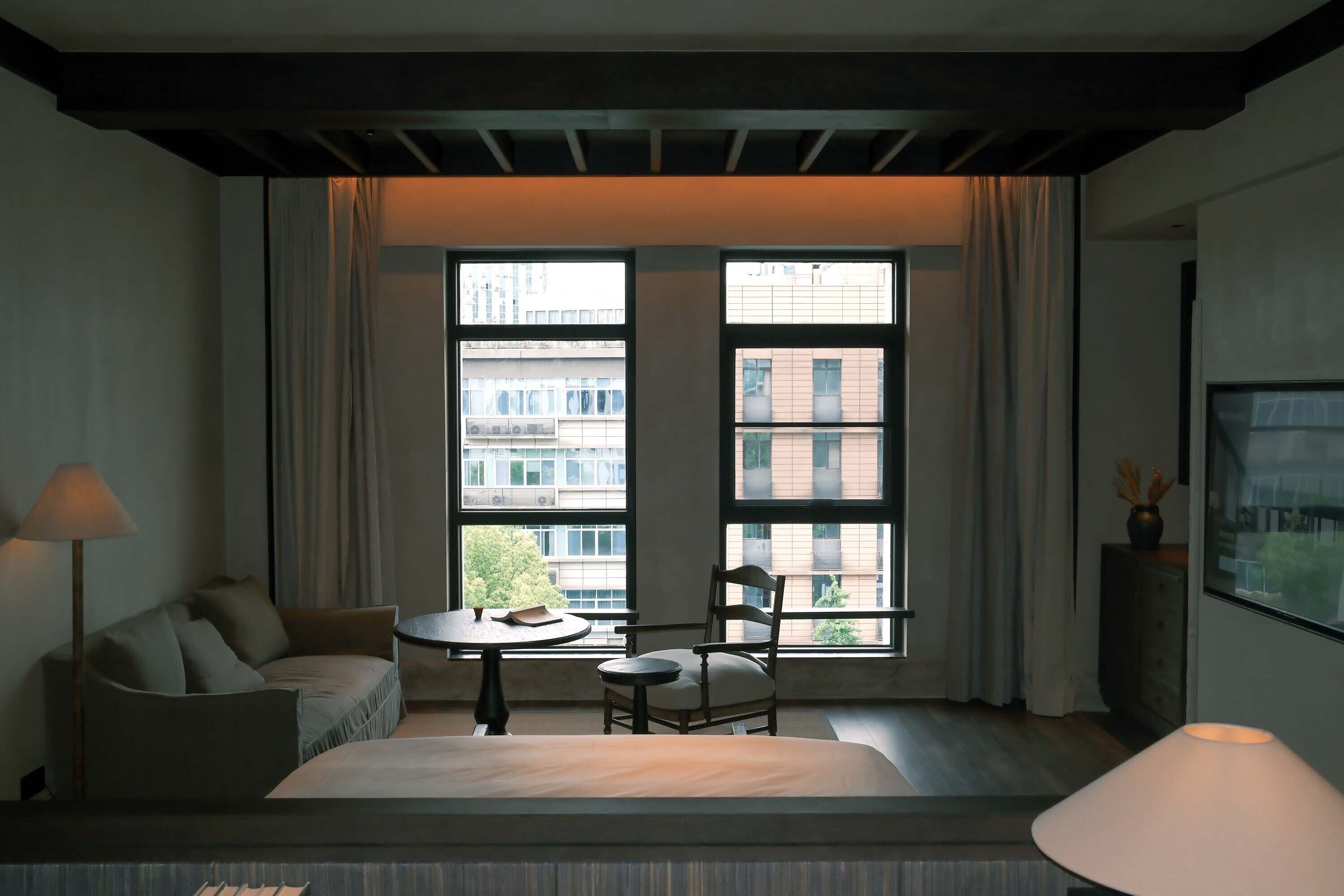 Habitación de hotel con cama, una silla, una mesita y una lámpara. Hay dos ventanas grandes con cortinas y vistas a edificios.