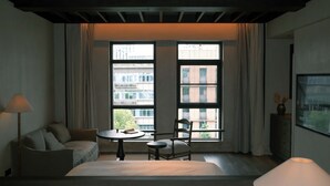 A hotel room with a bed, a chair, a small table, and a lamp. There are two large windows with curtains, and a view of buildings outside.