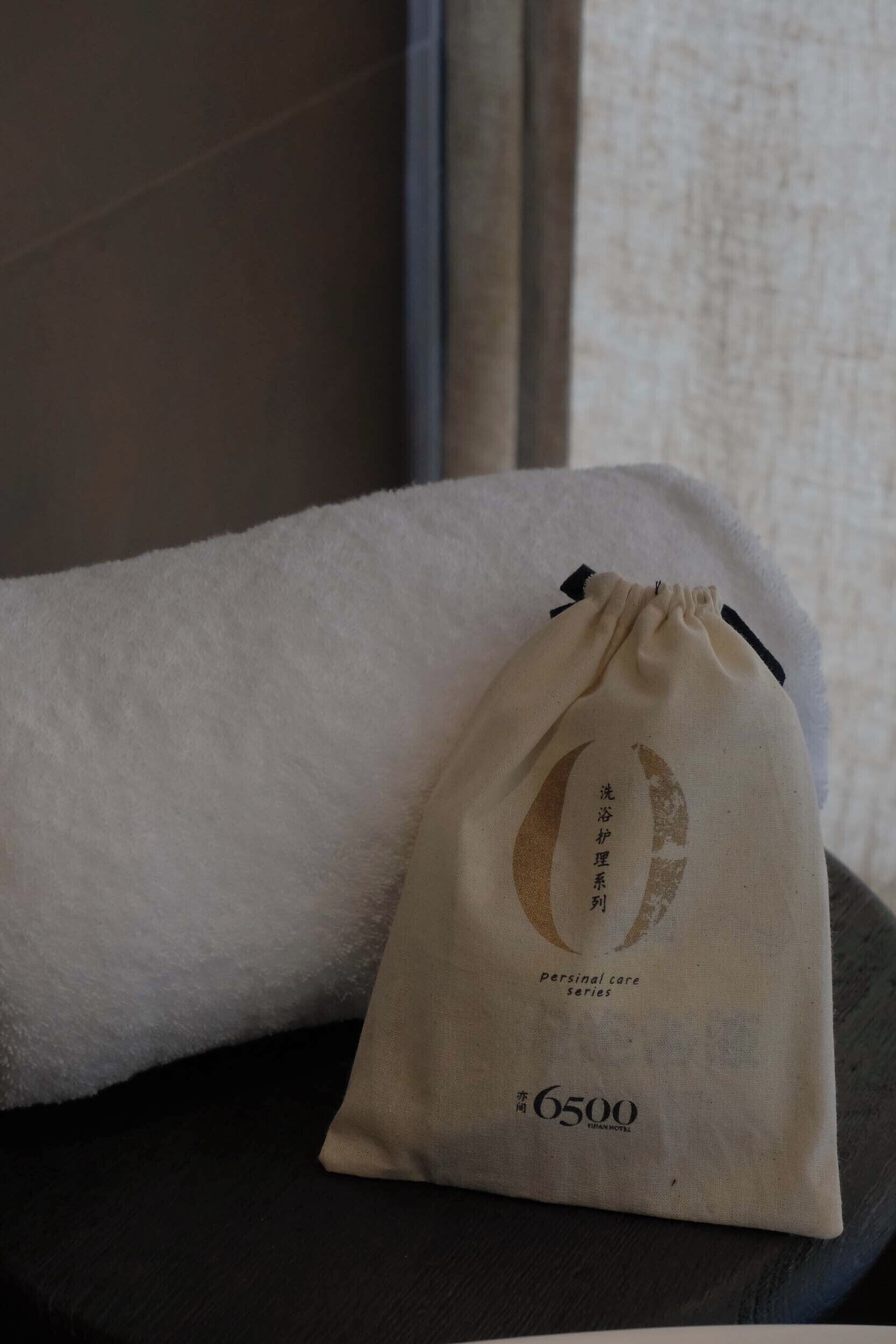 A small beige drawstring bag with a logo and text on it, placed on a dark surface next to a white towel.