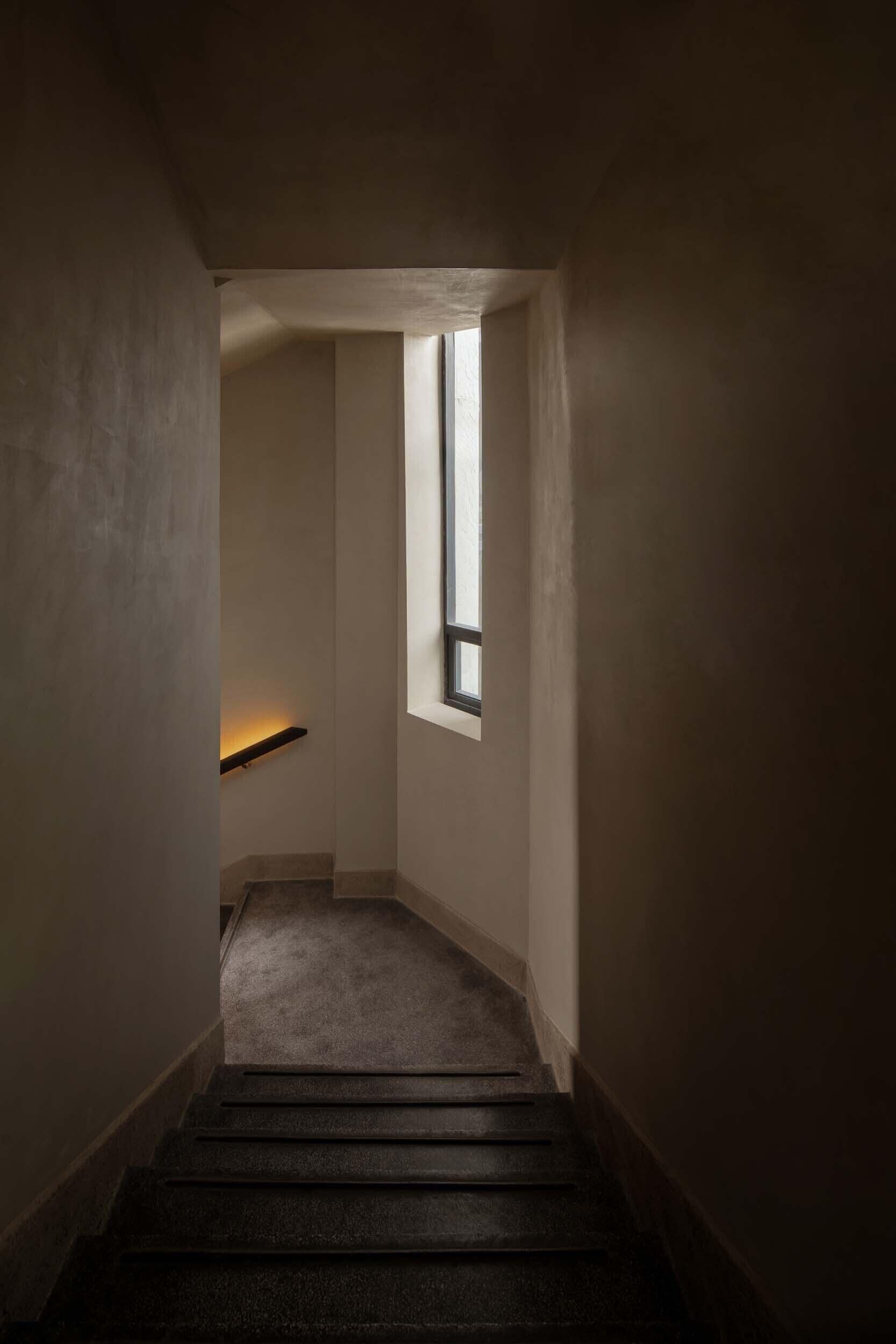 A narrow stairway leading to a landing with a window, a wall-mounted shelf, and a textured floor.