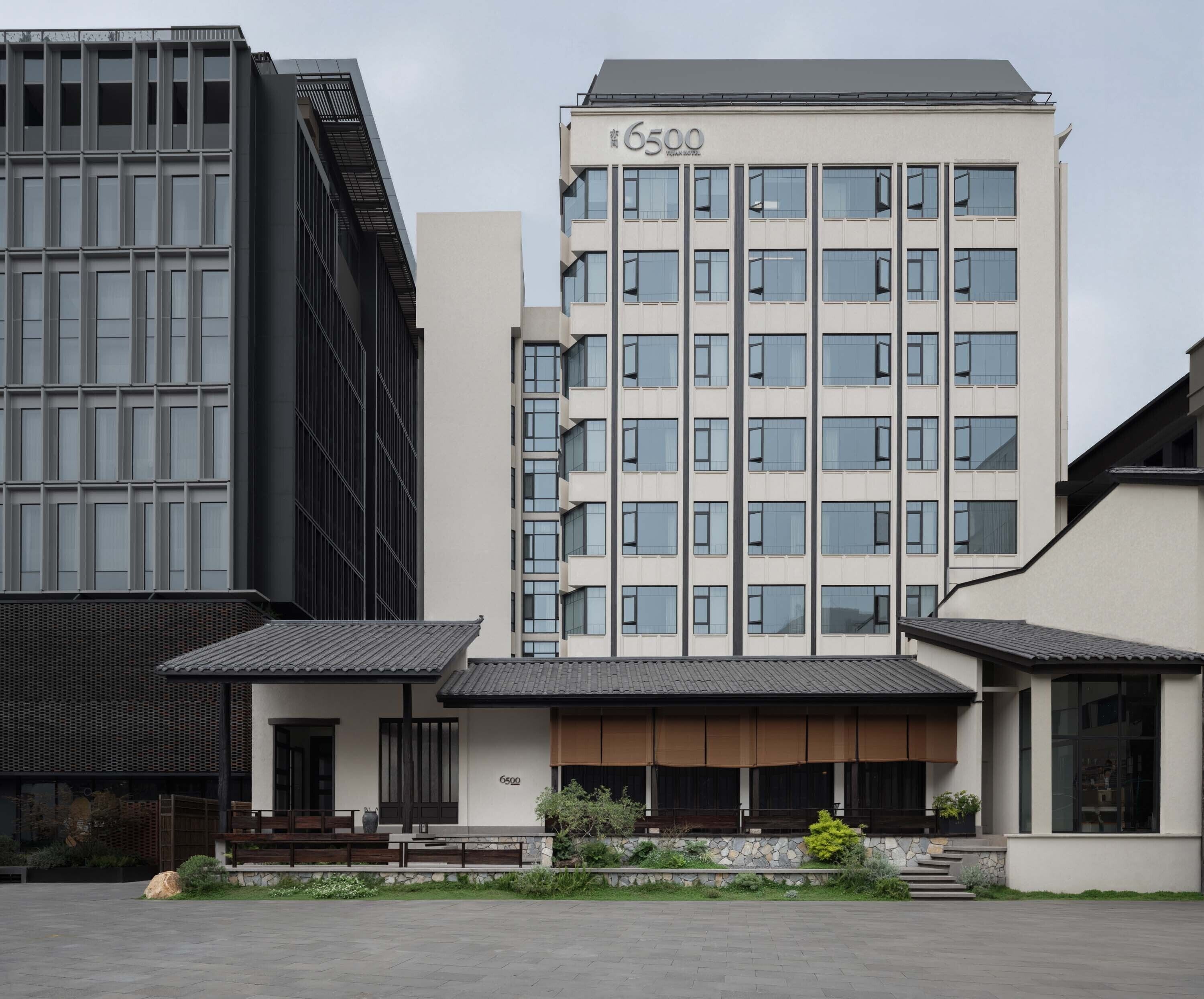 A modern hotel building with a prominent '6500' sign, featuring a mix of glass and concrete facade, and a traditional-style entrance with a stone pathway and greenery.