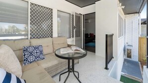 Ein Zimmer mit einer Couch, einem kleinen runden Tisch mit einem Buch, einem Fenster mit einem Fliegengitter und Blick ins Freie.