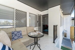 Ein Zimmer mit einer Couch, einem kleinen runden Tisch mit einem Buch, einem Fenster mit einem Fliegengitter und Blick ins Freie.