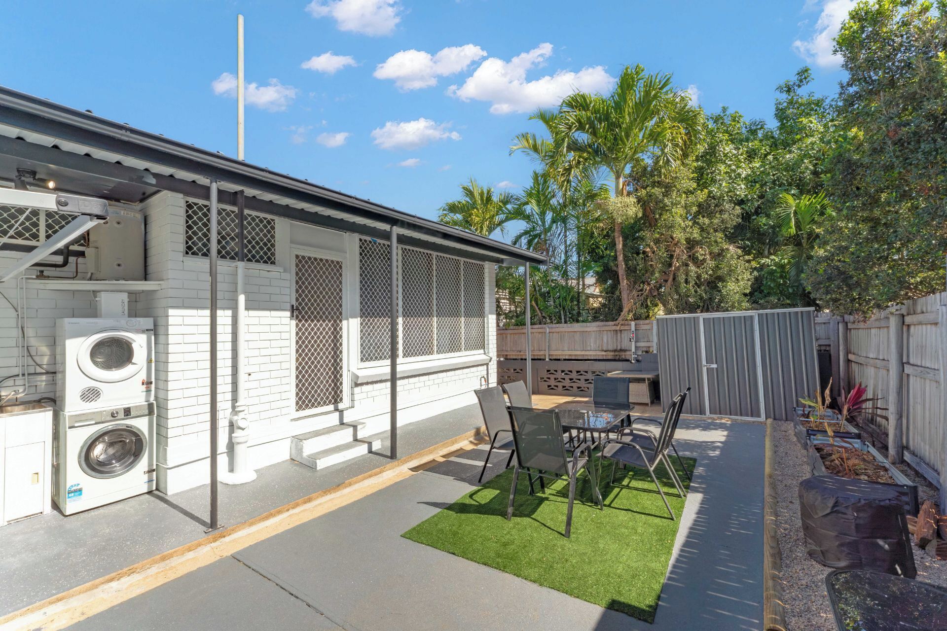 A residential backyard with a patio, outdoor furniture, a washing machine, and a fenced area with trees.