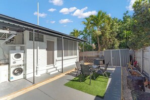 Un jardin à l’arrière d’une maison, comprenant une terrasse, du mobilier d’extérieur, un lave-linge et une zone délimitée par une clôture, avec des arbres.