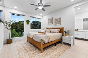 Une chambre moderne avec un grand lit, un ventilateur de plafond, une vue sur la verdure et une salle de bain avec un miroir.