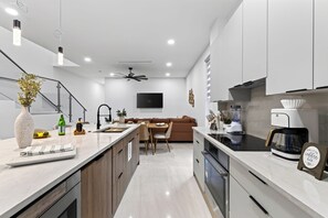 A modern kitchen with a central island, built-in appliances, and a dining area with a brown sofa. - Private Pool + Grill + Twin Homes | Victoria Park | Palm Perch + Palm Court (Fort Lauderdale)