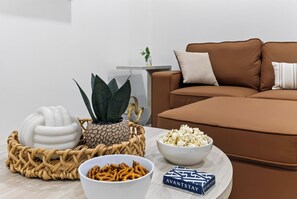 A modern living room with a brown sofa, a white bowl of popcorn, a bowl of pretzels, a decorative plant, and a woven basket. - Private Pool + Grill + Twin Homes | Victoria Park | Palm Perch + Palm Court (Fort Lauderdale)