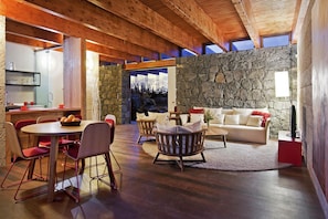 A modern interior with a stone wall, wooden ceiling beams, a dining area with red chairs, and a sofa with cushions. - Casas Bioclimáticas Iter La Compacta (Granadilla de Abona)