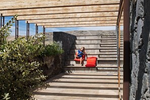 A wooden staircase leading to a stone wall with a person sitting on a red cushion. - Casas Bioclimáticas Iter La Compacta (Granadilla de Abona)