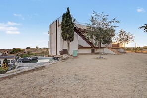 Un edificio moderno con una escalera que lleva a un segundo piso, rodeado de un patio arenoso con un árbol y una maceta grande.