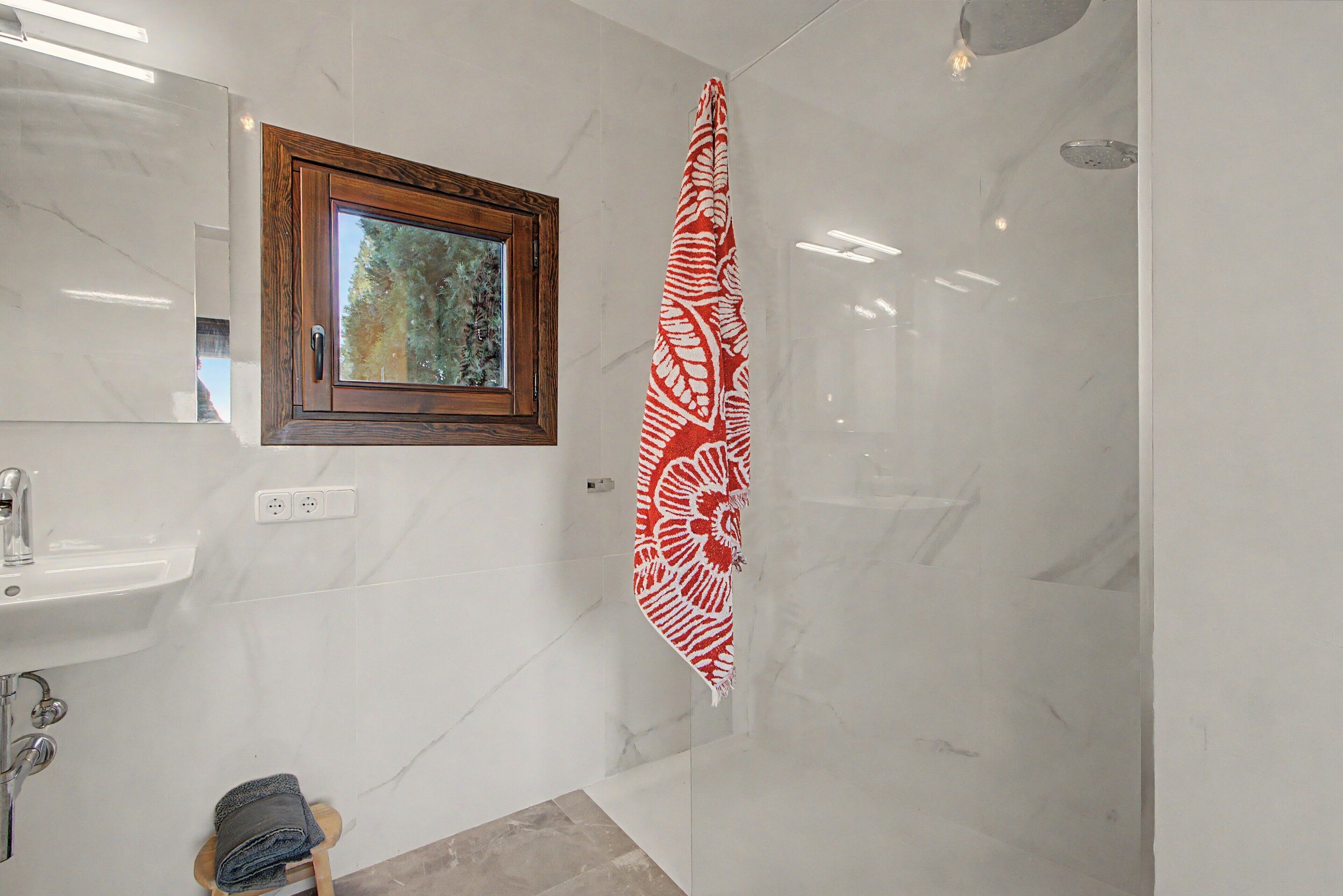 Un bagno con un lavandino bianco, uno specchio con cornice in legno, un portasciugamani con un asciugamano rosso decorato e una zona doccia.