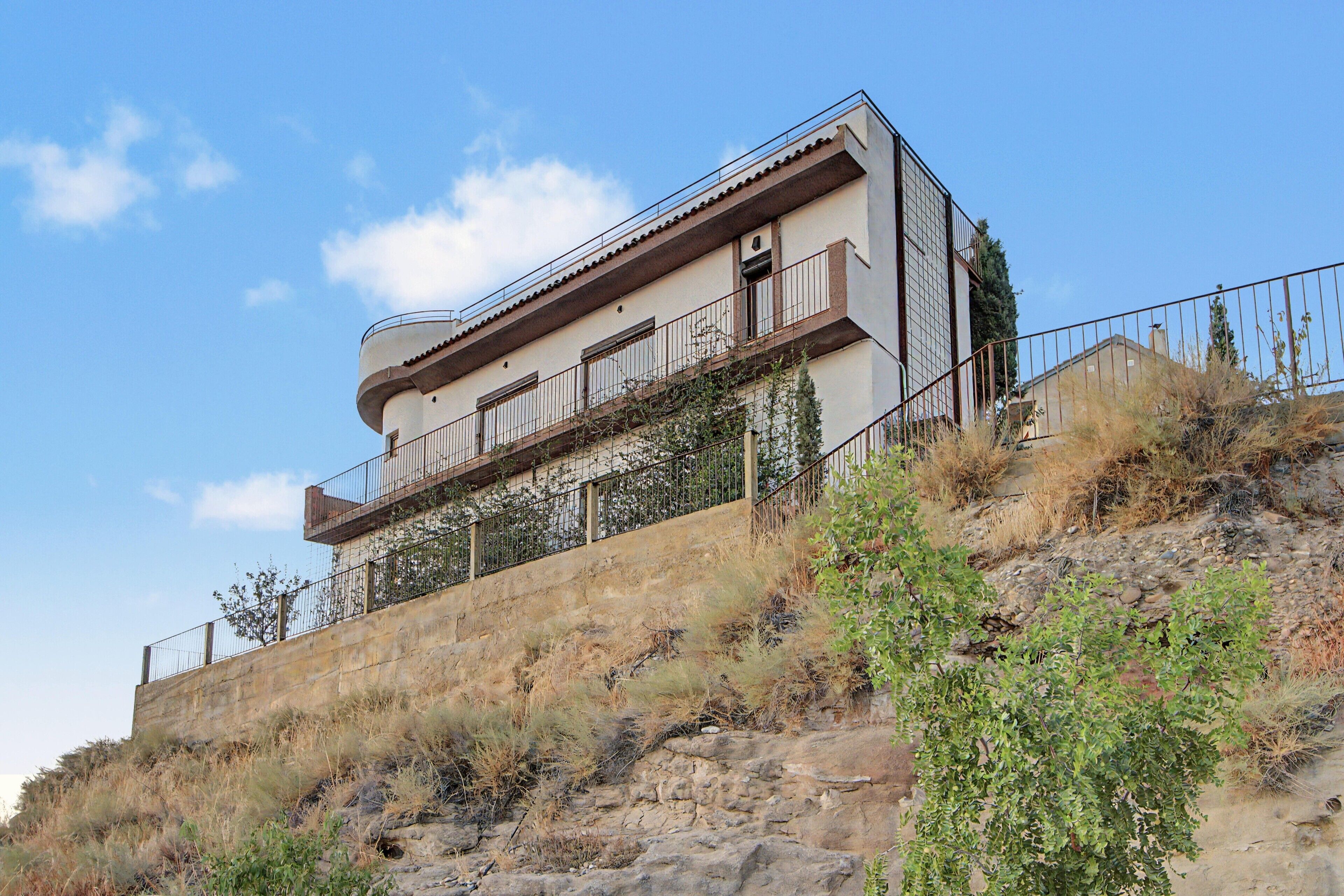 A multi-story building with balconies, situated on a hillside with sparse vegetation.