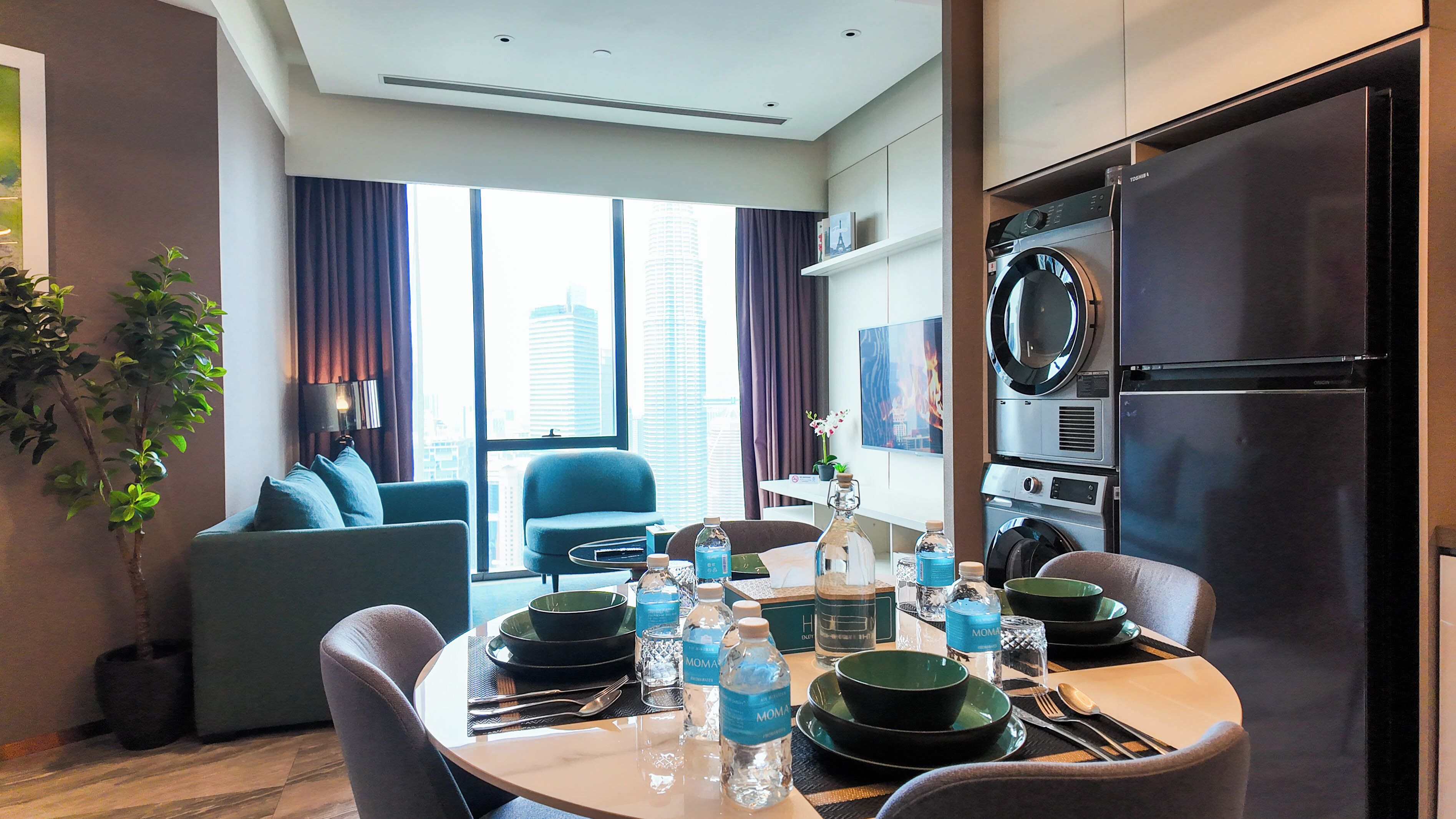 A modern hotel room with a dining area, a sofa, a TV, and a built-in washer and dryer.