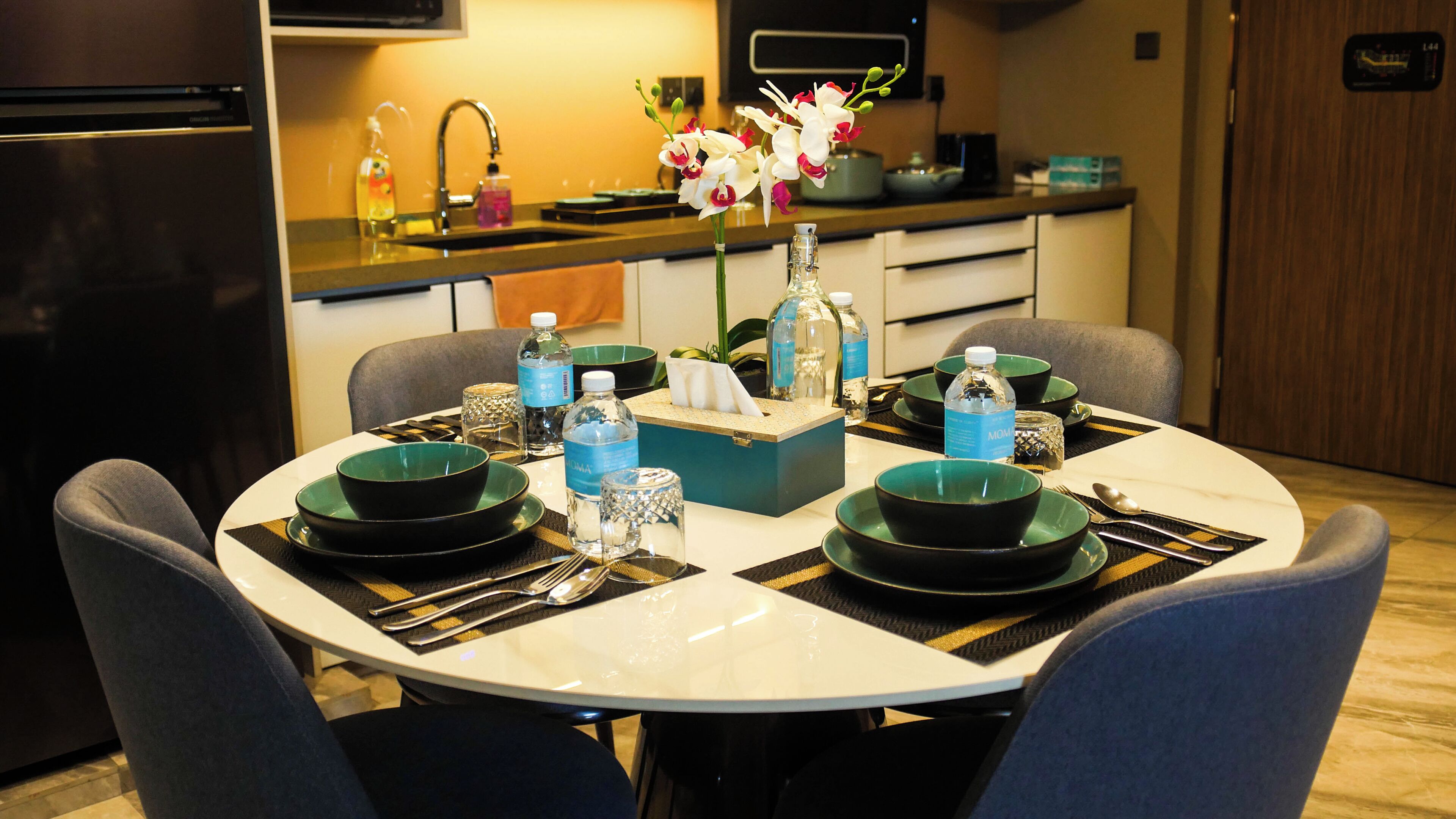 A dining area with a round table set for four, equipped with black plates, clear glassware, and a vase with flowers.