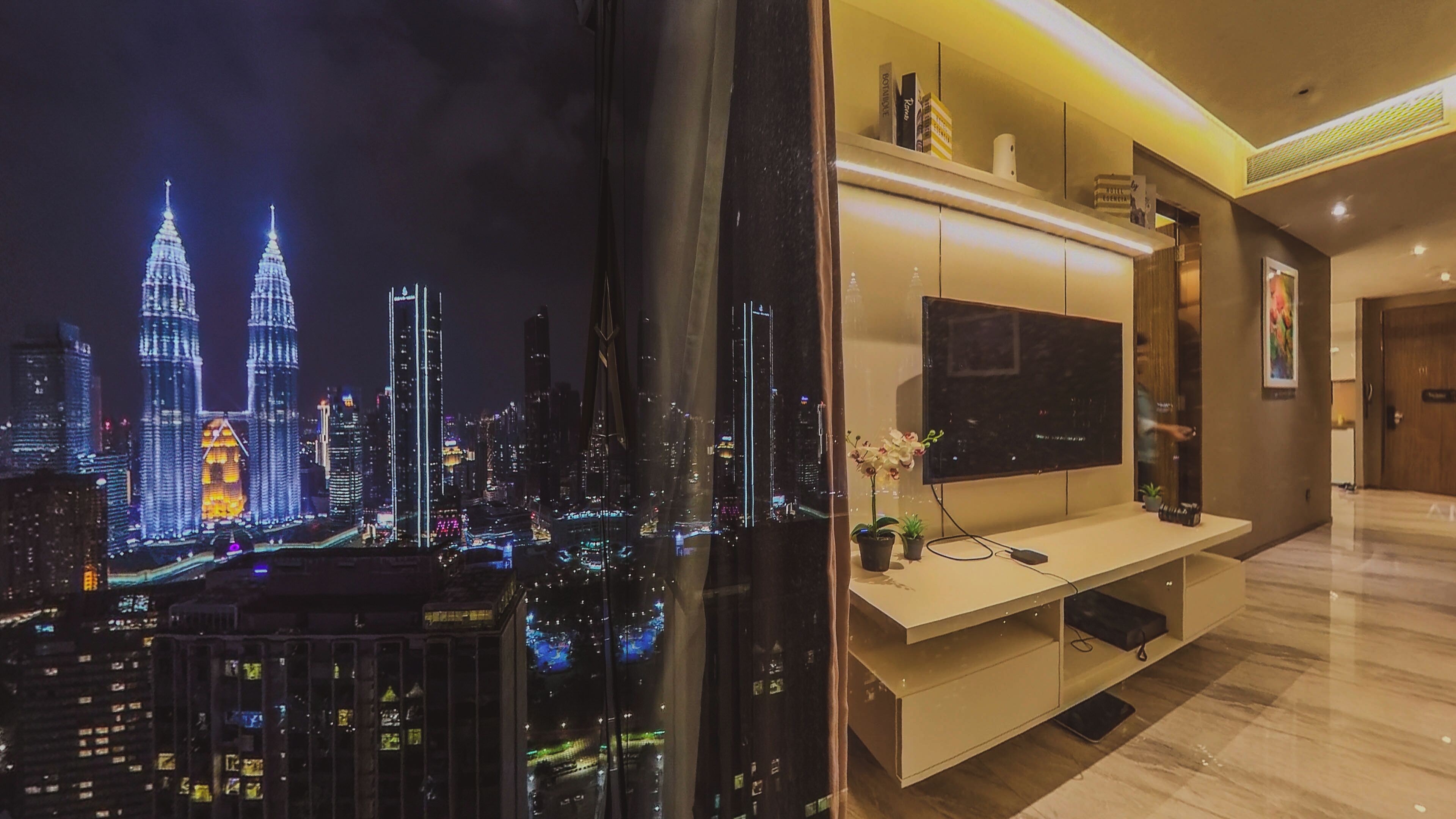 A modern living room with a large flat-screen TV mounted on a white cabinet, a glass wall with a cityscape view, and a small potted plant on the cabinet.