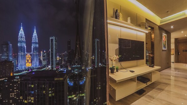 Een moderne woonkamer met een grote flatscreen televisie bevestigd aan een wit meubelstuk, een glazen wand met uitzicht op een stadsgezicht, en een kleine potplant op het meubelstuk.