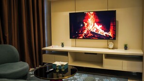 A living room with a flat-screen TV mounted on the wall, a sofa, a coffee table with remote controls, and a small clock. - Imperial Residence KLCC- JD Home (Wilayah Persekutuan)