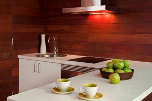 A modern kitchen with a stainless steel sink, white cabinets, and a white countertop. There are two white cups with green saucers and a basket of green apples. - Casas Bioclimáticas Iter El Muro (Granadilla de Abona)