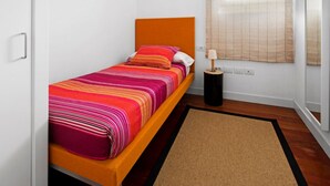 En säng för en person med en orange sänggavel, ett färgglatt randigt överkast och ett litet nattduksbord med en lampa.